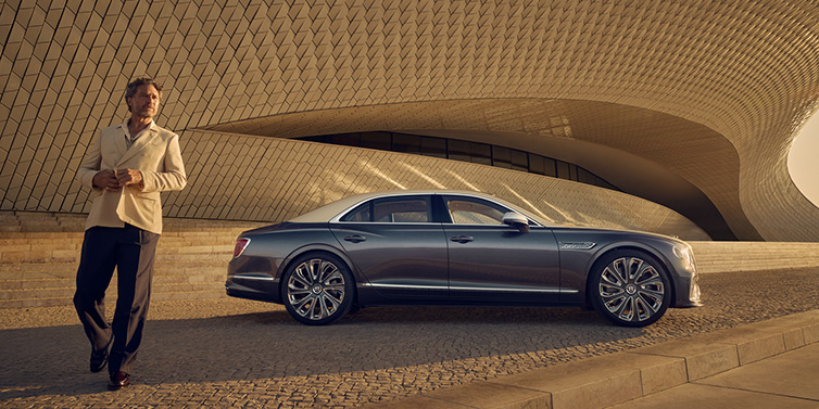 Bentley Brisbane Bentley Flying Spur Mulliner sedan in Rose Gold over Magnetic duotone paint parked by a curved wall with person in suit jacket standing on the left